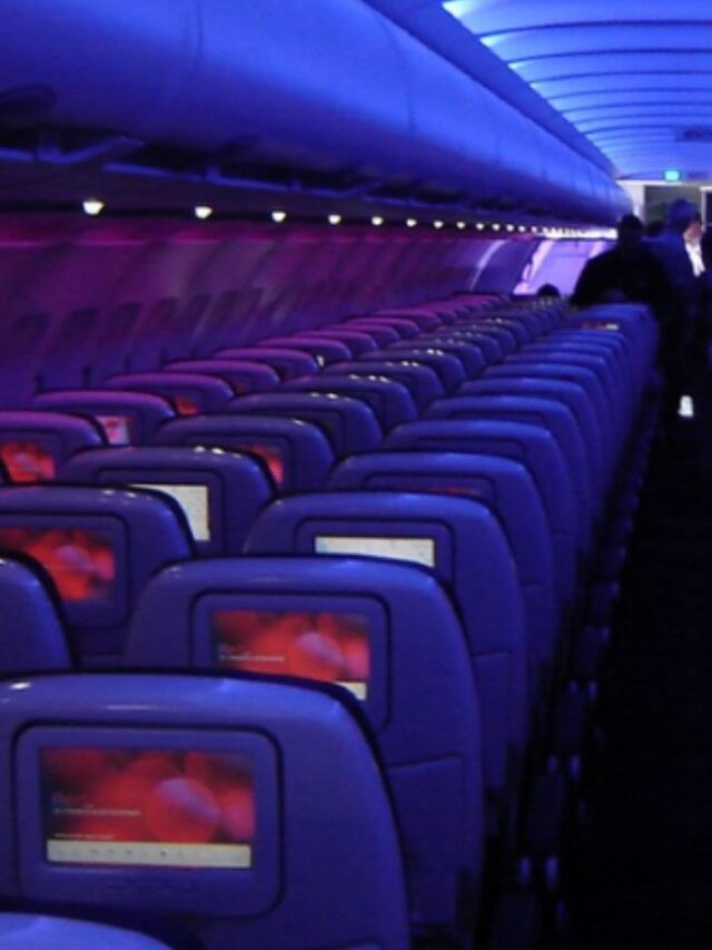Airplane cabin with rows of economy seats, each equipped with seatback screens, illuminated by purple and blue ambient lighting.